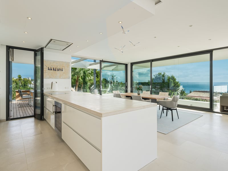 Cuisine blanche moderne avec grand îlot, salle à manger et baies vitrées du sol au plafond donnant sur l'océan.