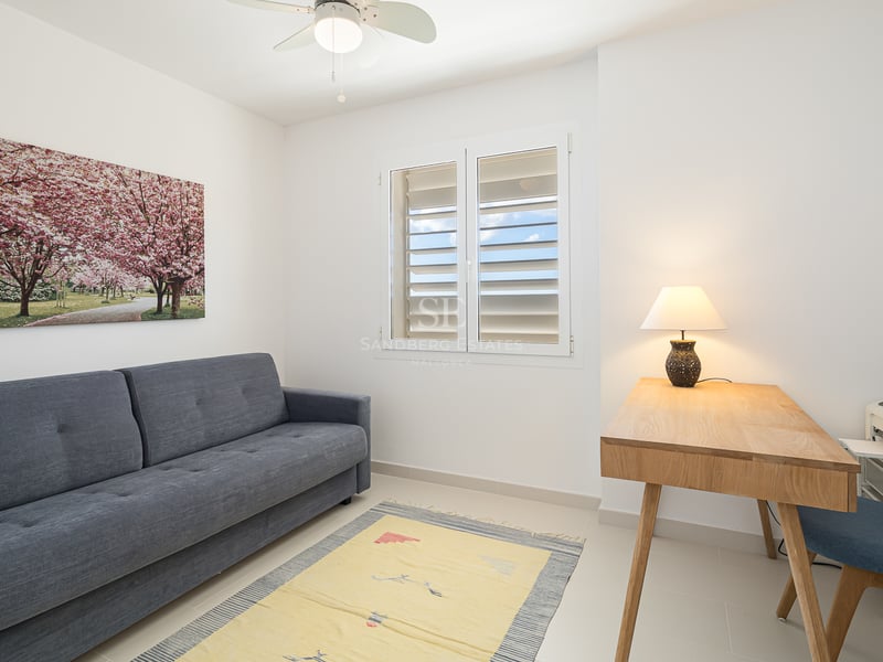 Pièce minimaliste avec un canapé gris, un bureau en bois avec une lampe, une chaise bleue et une fenêtre avec volets blancs.