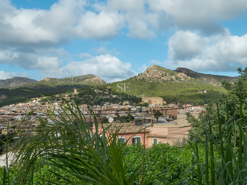 Vue d'un village méditerranéen aux toits de terre cuite entouré de montagnes verdoyantes sous un ciel nuageux.