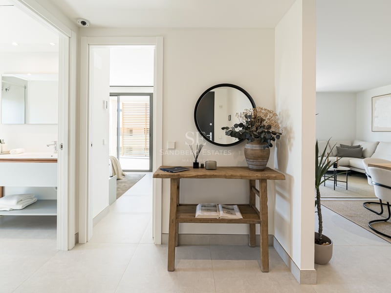 Pasillo luminoso con consola de madera rústica, espejo redondo y vistas al baño y salón de diseño moderno.