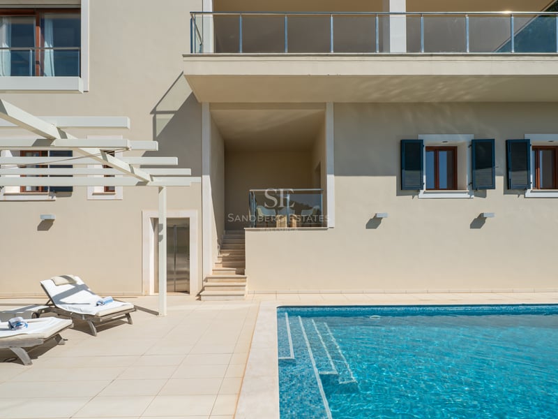 Piscine de villa de luxe avec mosaïque et chaises longues blanches sur une terrasse en pierre sous une pergola.