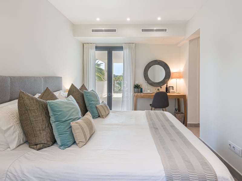 Chambre lumineuse avec grand lit, coussins turquoise, bureau en bois avec miroir rond et porte vitrée vers la terrasse.