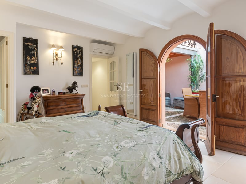 Chambre avec portes en bois voûtées ouvrant sur un patio ensoleillé, comprenant une salle de bain avec mur en pierre.