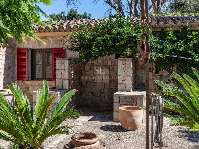 Espace de douche extérieure en pierre avec volets rouges, pots en terre cuite et plantes vertes dans une cour méditerranéenne.