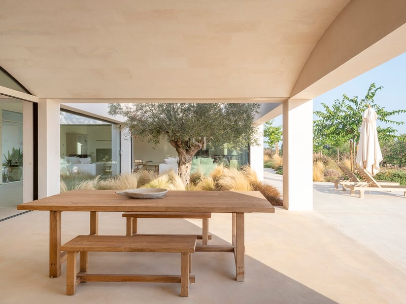A wooden dining table on a covered sandstone terrace next to an olive tree and sun loungers.