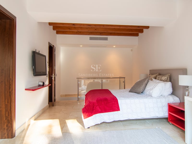 Chambre moderne avec lit double, touches de rouge, poutres en bois au plafond et sol en pierre crème.