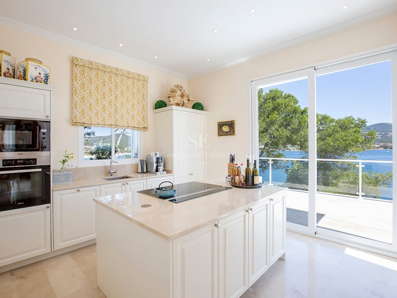 Cuisine moderne crème avec îlot central et grandes baies vitrées donnant sur la mer et des pins.
