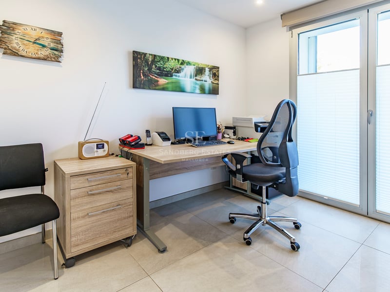 Bureau avec bureau en bois, chaise ergonomique, ordinateur et grande porte-fenêtre avec stores.