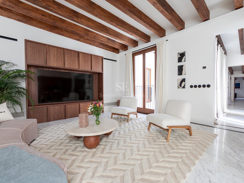 Salón luminoso con vigas de madera oscura, suelo de mármol, sillones bouclé blancos y mueble de televisión a medida.