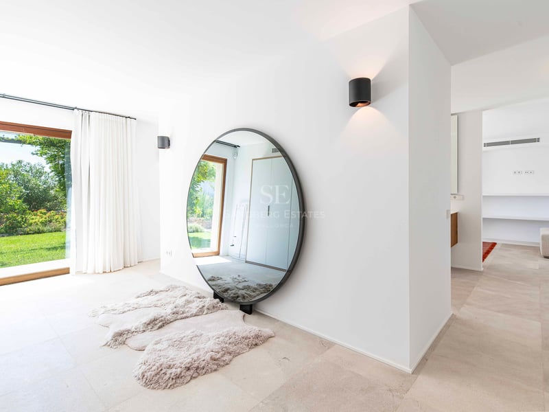 Intérieur minimaliste lumineux avec un grand miroir rond, sol en pierre et baies vitrées ouvrant sur un jardin verdoyant.