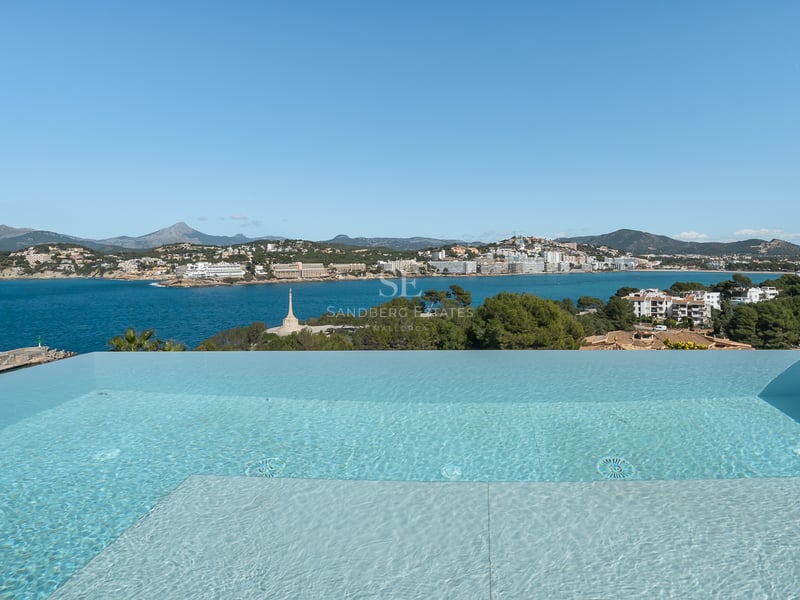 Une piscine à débordement surplombant une baie bleue, des bâtiments côtiers et des montagnes.