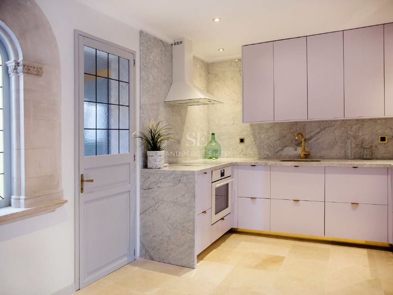 Cocina contemporánea con armarios blancos, salpicadero de mármol, grifo dorado y una ventana de piedra arqueada.