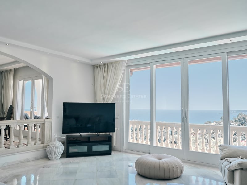 Salon lumineux avec sol en marbre blanc et grandes baies vitrées ouvrant sur une terrasse avec vue sur la mer.