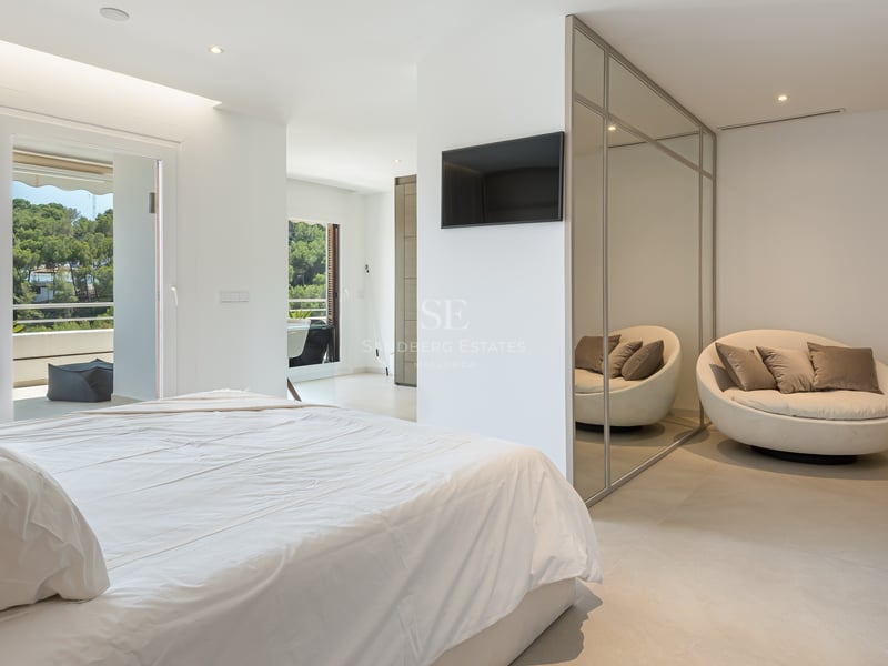 Chambre lumineuse avec lit blanc, armoire en miroir, fauteuil circulaire et vue sur la terrasse et les arbres.