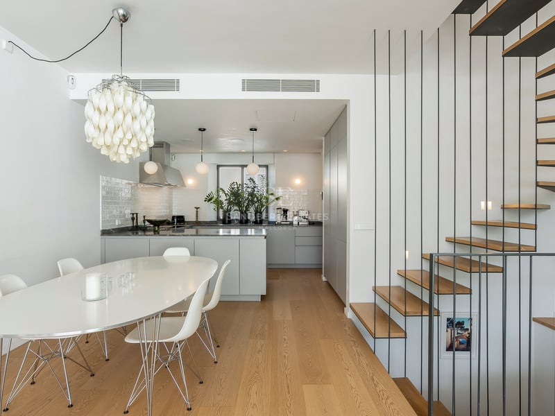 Salle à manger blanche avec table ovale, chaises Eames, lustre design, cuisine grise et escalier en bois et métal.