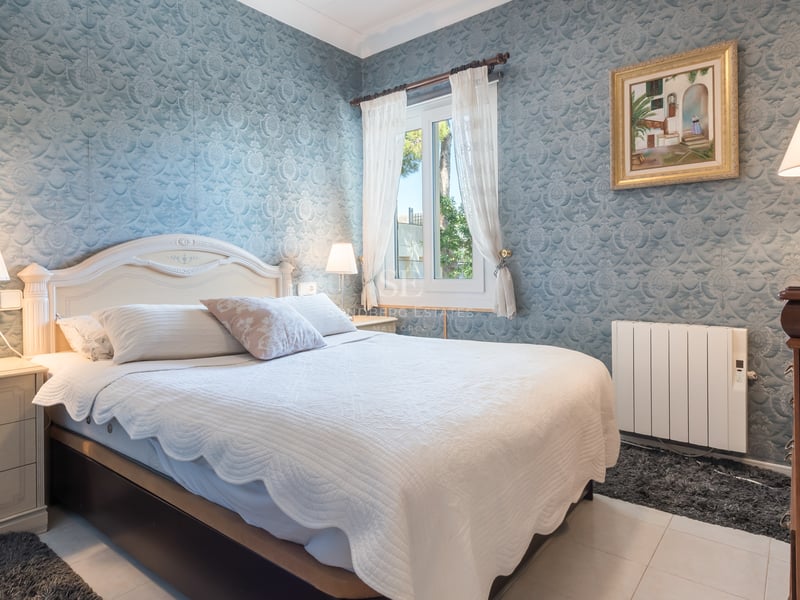 Chambre de style classique avec papier peint bleu à motifs, lit blanc et tables de chevet en bois sous une lumière douce.