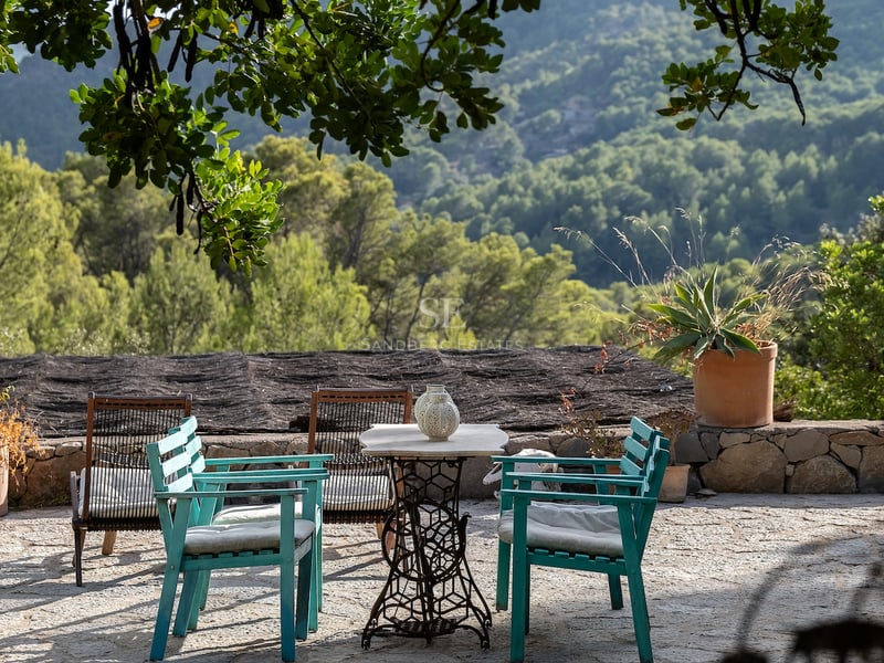 Terrasse en pierre avec chaises turquoise et table vintage surplombant une forêt verdoyante et des montagnes.