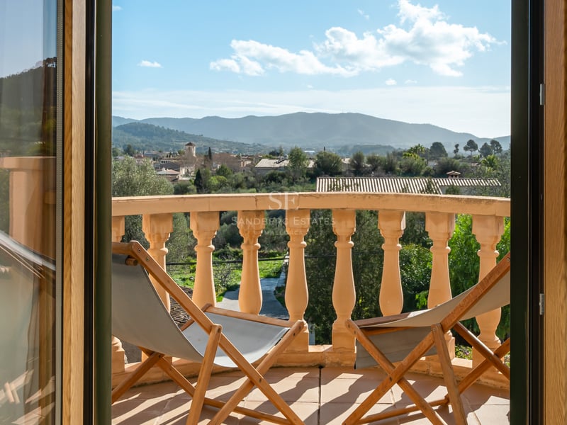Balcon ensoleillé avec balustrade en pierre, deux chaises longues en bois et vue sur un village et les montagnes.