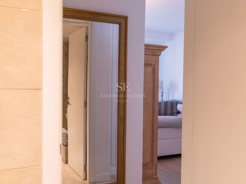 Vue à travers une porte ouverte montrant un couloir avec un miroir doré et une chambre avec armoire en bois.
