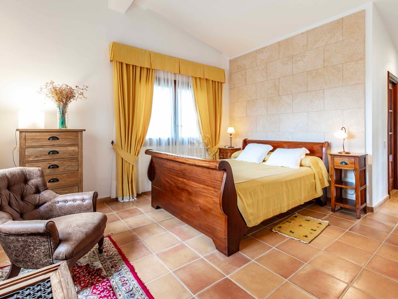 Une chambre rustique avec un lit en bois, des tomettes en terre cuite, un fauteuil en cuir et des rideaux jaunes.