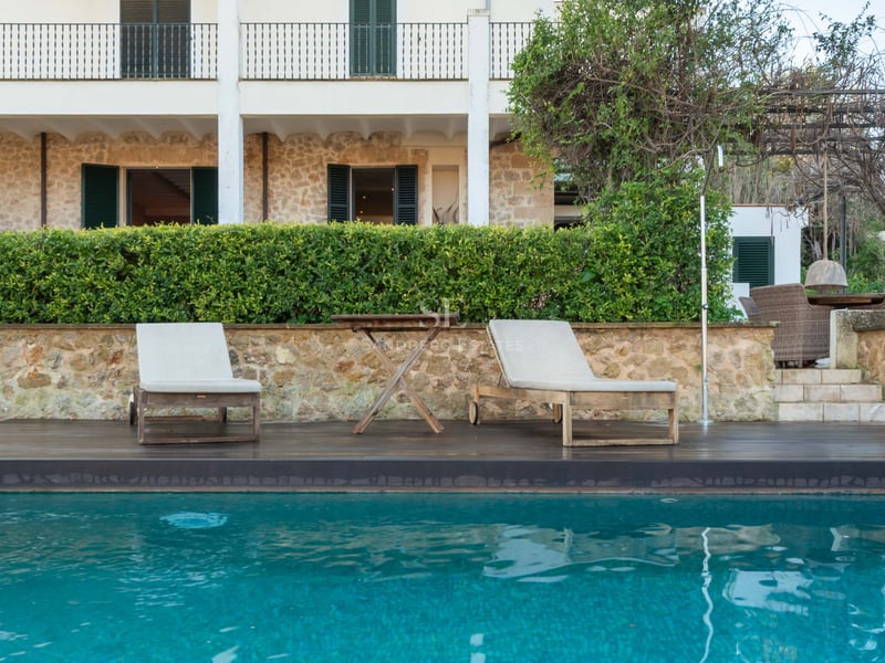 Piscine turquoise avec chaises longues en bois devant une villa en pierre aux volets sombres.
