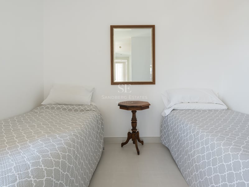 Chambre blanche avec deux lits simples, couvre-lits gris à motifs et une table d'appoint ancienne en bois.