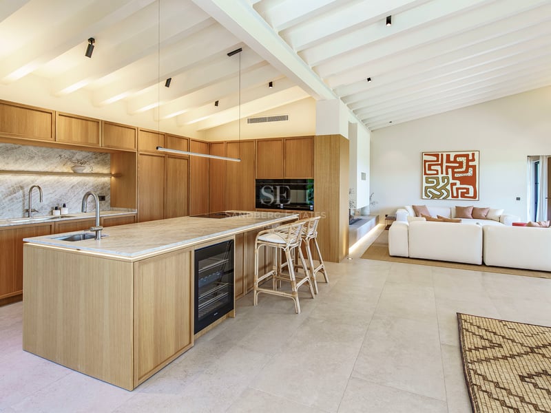 Cocina moderna de planta abierta con muebles de madera clara, isla de mármol y techo de vigas blancas.