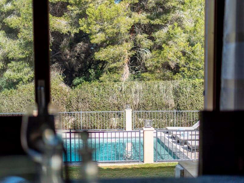 Vue par une fenêtre d'une piscine turquoise clôturée entourée de transats et de grands pins.