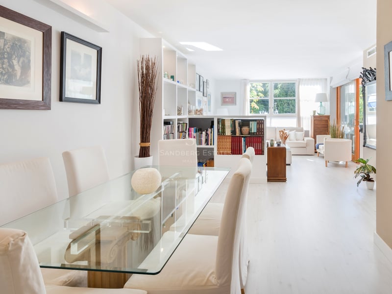 Salle à manger ouverte avec table en verre et chaises blanches menant à un salon lumineux avec bibliothèque.