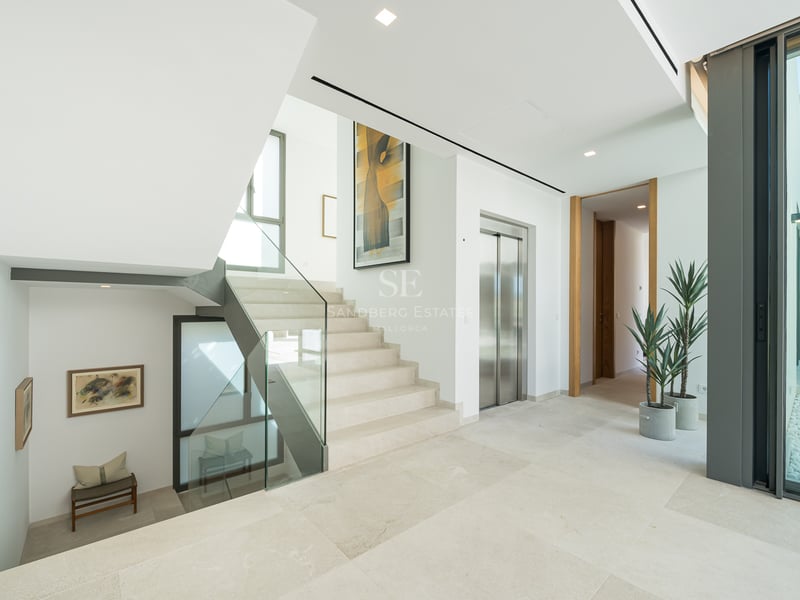 Hall moderne lumineux avec un escalier en pierre claire, garde-corps en verre, un ascenseur en inox et de grandes fenêtres.