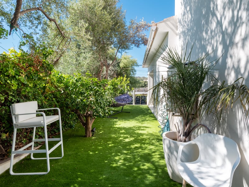 Petit jardin latéral avec gazon synthétique, chaises design blanches, arbres verdoyants et mur de villa blanc.
