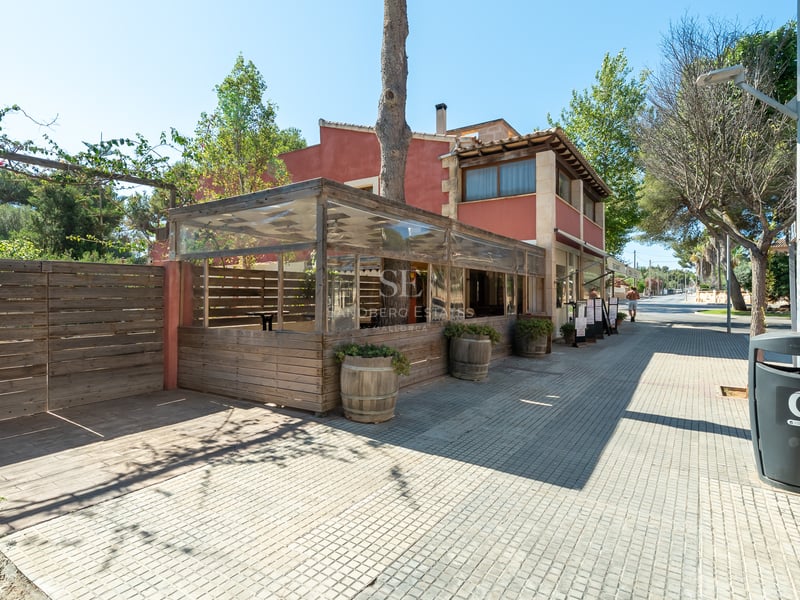 Façade rouge terre cuite avec terrasse en bois fermée sur un trottoir ensoleillé à Calvià.