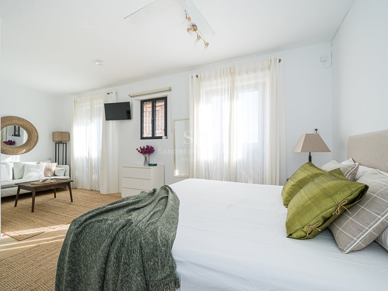 Chambre principale lumineuse avec un grand lit, coussins verts, un coin salon avec canapé blanc et tapis en jute.