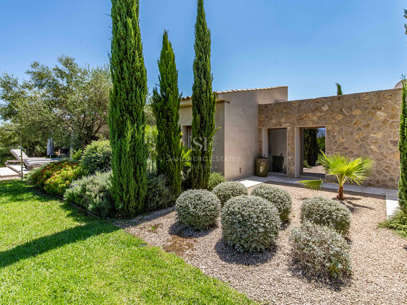 Villa moderne aux murs en pierre naturelle et jardin avec cyprès et arbustes sous un ciel bleu clair.