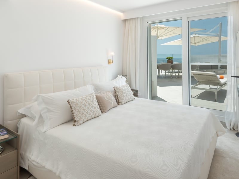 Chambre principale lumineuse avec tête de lit capitonnée et baies vitrées ouvrant sur une terrasse face à la mer.
