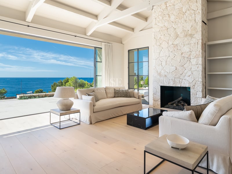 Salon lumineux avec cheminée en pierre, canapés crème et baies vitrées ouvrant sur la Méditerranée.