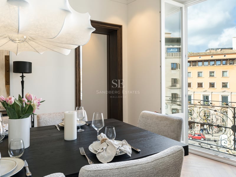 Modern dark wood dining table and white chairs facing an open balcony overlooking historic city architecture.