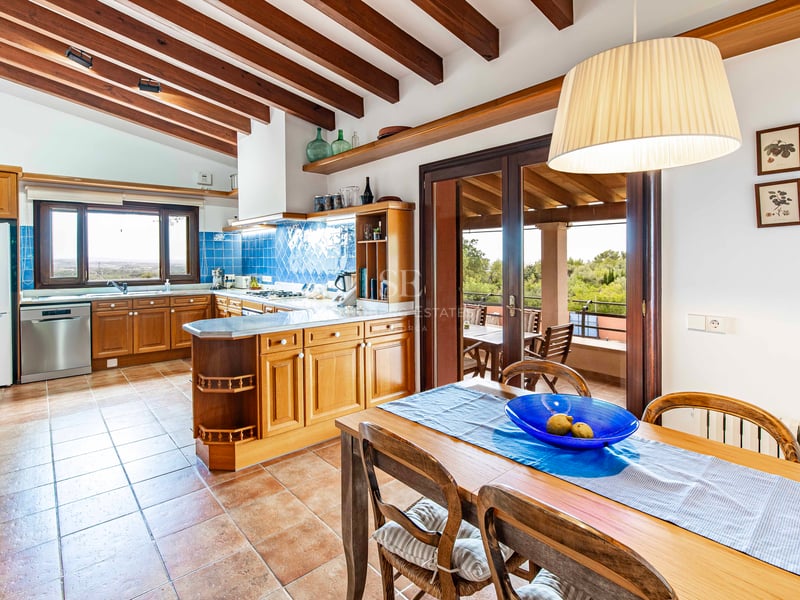 Cuisine avec meubles en bois, carrelage bleu, sol en terre cuite et poutres apparentes avec table à manger.