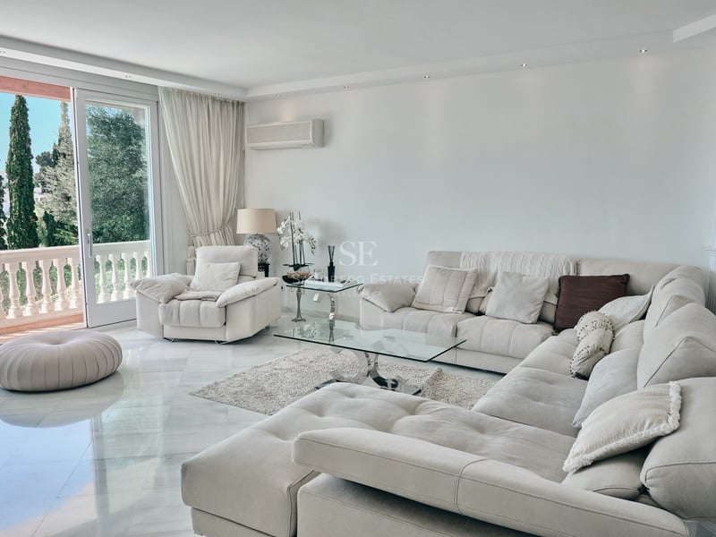 Salon spacieux avec canapé d'angle beige, sol en marbre blanc et vue sur jardin méditerranéen.