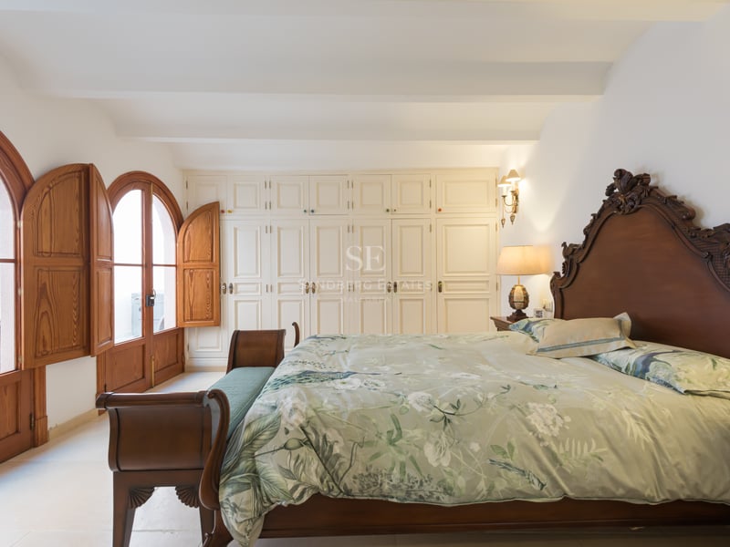 Grande chambre avec lit en bois sculpté, literie florale, placards blancs et portes cintrées en bois avec volets.