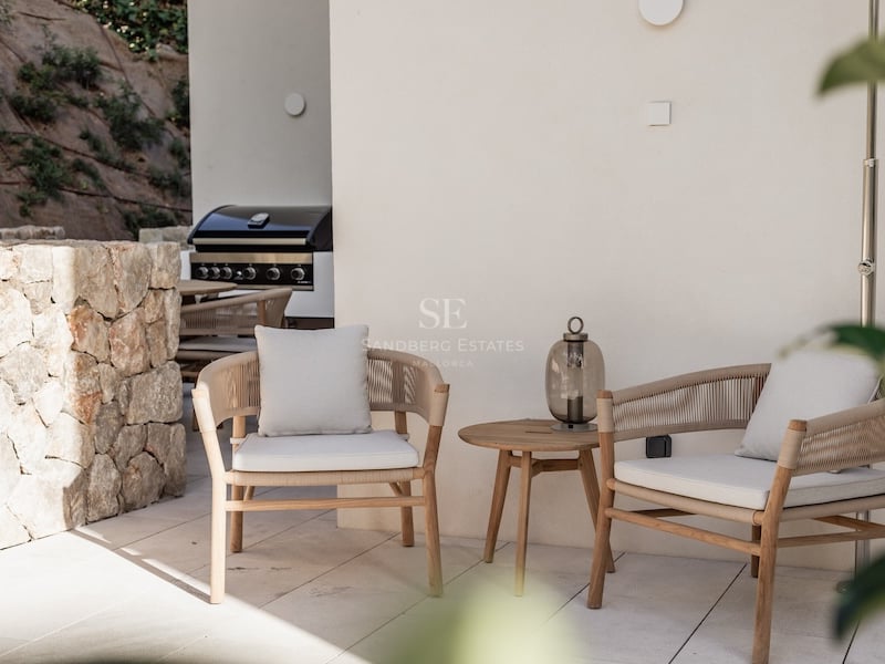 Deux élégants fauteuils modernes avec coussins à côté d'une table en bois sur une terrasse en pierre avec un barbecue.