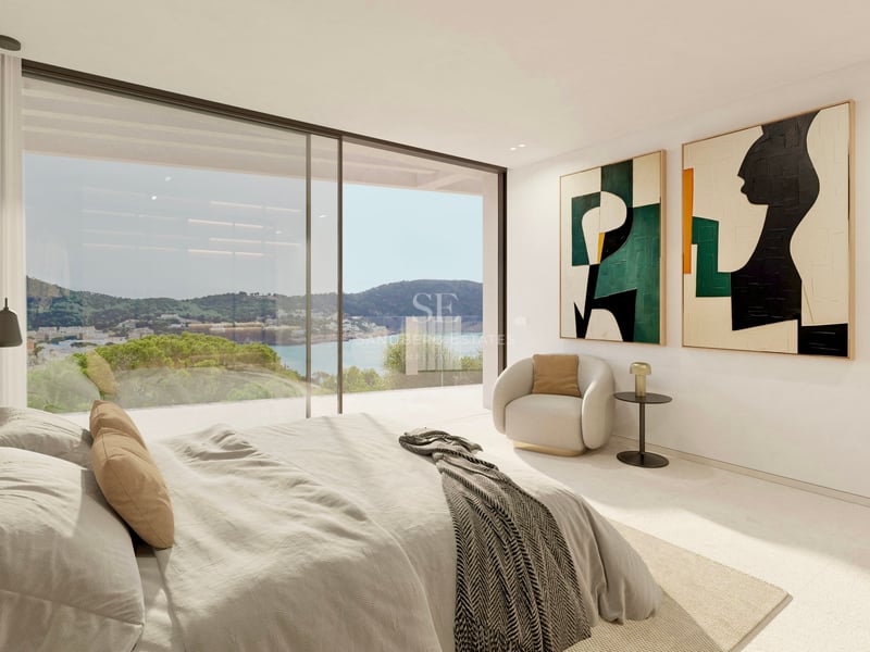 Chambre parentale moderne avec grandes baies vitrées donnant sur un paysage côtier, art abstrait et fauteuil.