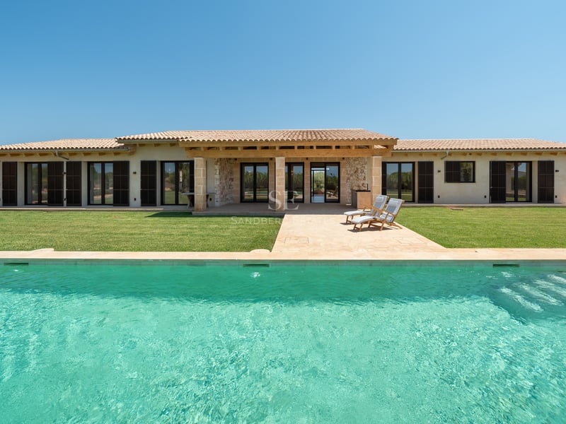 Piscina rectangular turquesa frente a una villa moderna de piedra de una planta con persianas oscuras y jardín.
