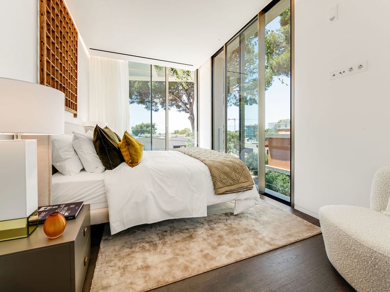 Chambre contemporaine lumineuse avec lit blanc, parquet foncé, fauteuil bouclé et parois vitrées.