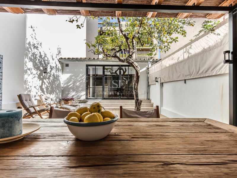 Terrasse ensoleillée avec table en bois, citronnier, chaises longues et murs blancs sous un toit en roseau.