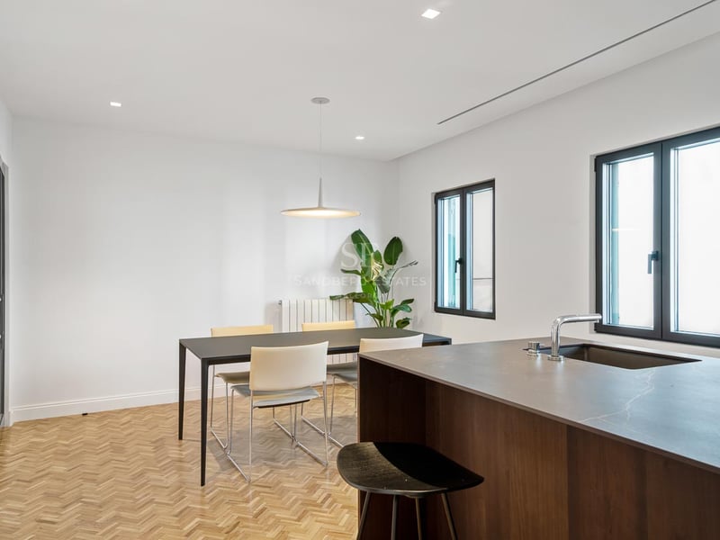 Salle à manger moderne avec table noire, chaises blanches, parquet en chevrons et îlot de cuisine.