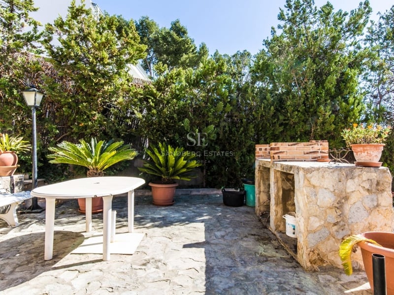 Patio pavé avec barbecue en pierre, table et banc blancs, entouré de haies vertes et de plantes en pots.