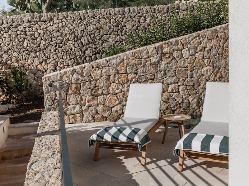 Deux chaises longues en bois avec coussins blancs devant un mur en pierre rustique sur une terrasse ensoleillée.