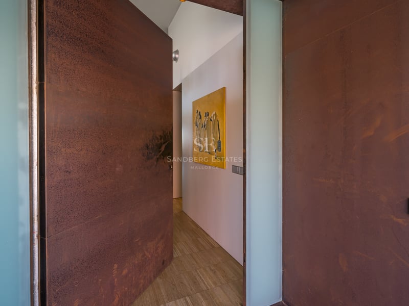 Grande porte en acier Corten s'ouvrant sur un couloir minimaliste avec murs blancs et sol en pierre.