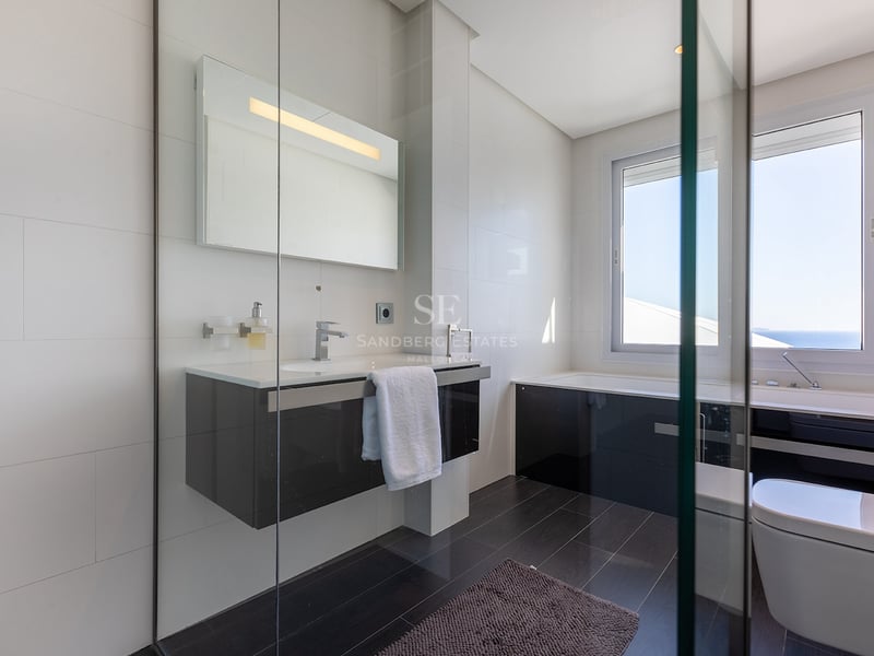 Salle de bain moderne avec murs carrelés blancs, sol sombre, vanité suspendue et baignoire sous une grande fenêtre.